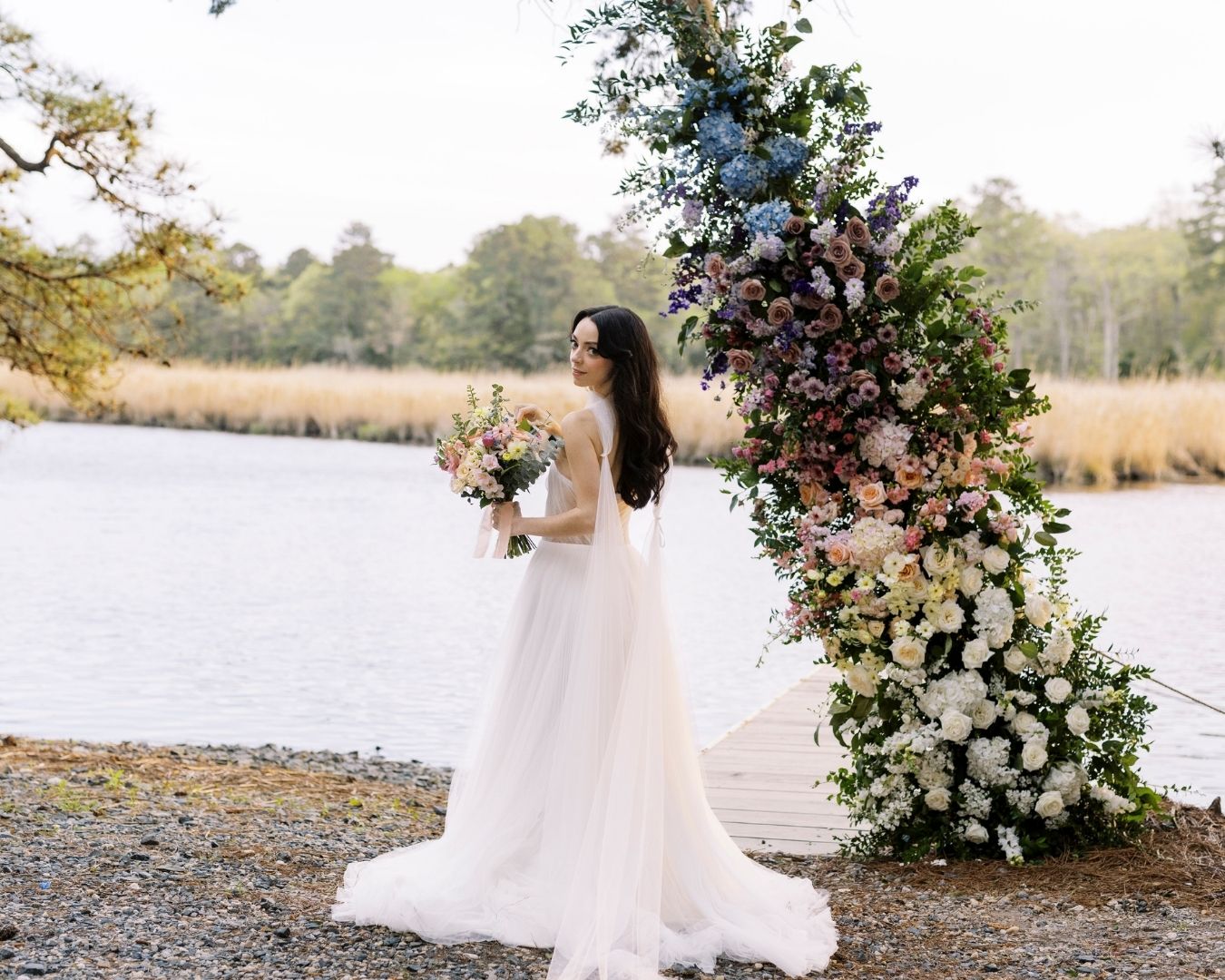 Bride looking over her shoulder while carrying a bouquet. She stands at the water's edge with a tree wrapped in colorful, romantic florals behind her.