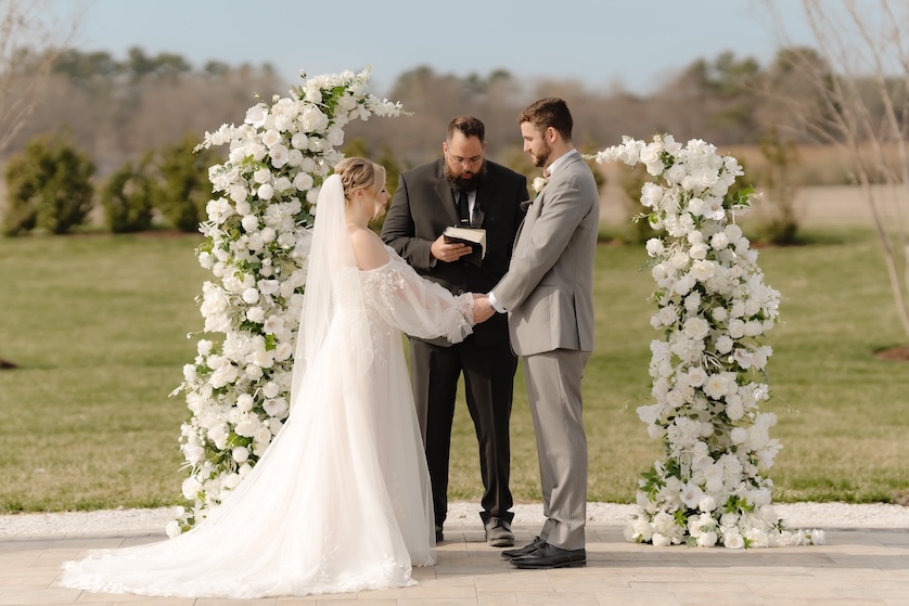 Bride and groom during ceremony in front of white pillar florals by Winters Farm Florals