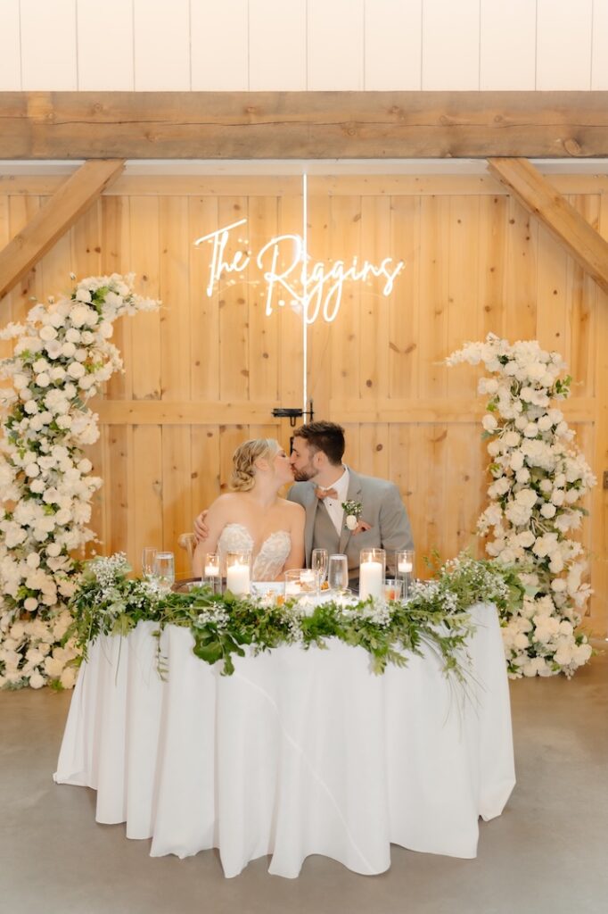 Bride and groom kissing at a sweetheart table adorned with greenery, with white floral pillars by Winters Farm Florals behind them