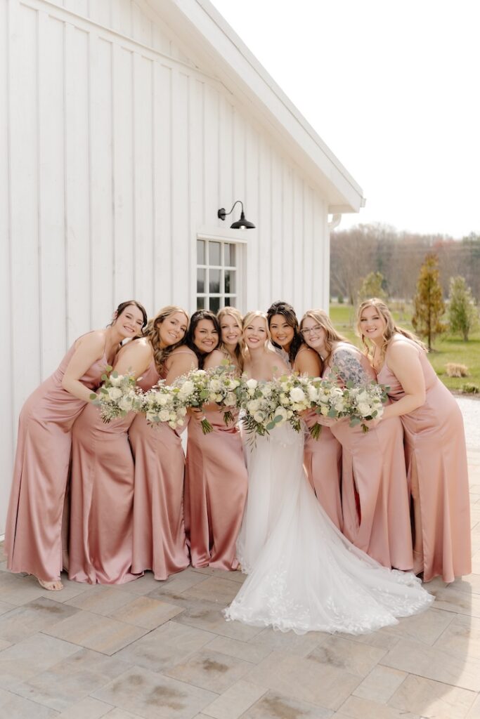 Bride and her bridesmaids in soft blush dresses all holding their green and white bouquets by Winters Farm Florals in front of them for the camera