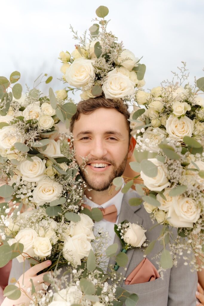 Groom smiling at the camera surrounded by soft whites and creams and greenery bouquets by Winters Farm Florals